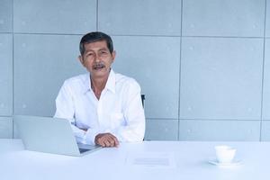 Work from home and social distancing concept. Asian business old man  working and drinking coffee at co-working space. Aging man using laptop meeting online. photo