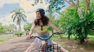 jovem latina no capacete protetor está andando de bicicleta ao longo da ciclovia em um parque da cidade plantado por árvores verdes. dia ensolarado. 4k cinematográfico