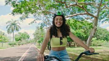 Young Latin woman in protective helmet is riding her bicycle along the bike path in a city park planted by green trees. Sunny day. Cinematic 4K video