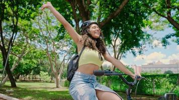 Young Latin woman in protective helmet is riding her bicycle along the bike path in a city park planted by green trees. Sunny day. Cinematic 4K video