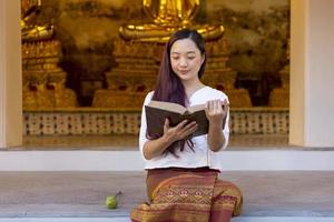 Asian buddhist woman is reading Sanskrit ancient Tripitaka book of Lord Buddha dhamma teaching while sitting in temple to chant and worship in the monastery photo