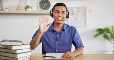 Portrait of Young Asian customer service support agent telemarketer wearing headset waving hand and looking at camera make business conference internet video call.