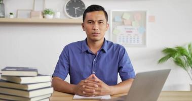 Portrait of young Asian man in blue shirt smiling and looking at camera while sitting at workdesk. video