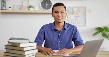 Portrait of young Asian man in blue shirt smiling and waving hand while sitting at workdesk. Male looking at camera. video