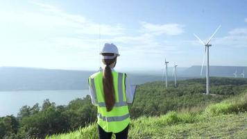 trabalhador de colarinho adulto ou maduro em capacete branco e uniforme verde desgaste permanente com documento contra usina de energia eólica na bela paisagem.
