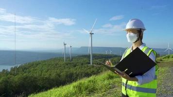 ingenieros asiáticos o mujeres técnicas con máscara y casco blanco están revisando el sistema de turbinas eólicas con un documento en una granja de energía renovable