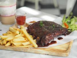 costillas de cerdo a la barbacoa, costillas de cerdo a la barbacoa con patatas fritas, ensalada de verduras, salsa de tomate en un vaso transparente en bandeja de madera, comida deliciosa foto