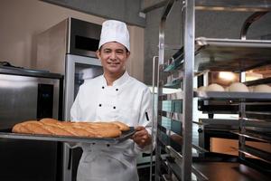 Senior Asian male chef in white cook uniform and hat showing tray of fresh tasty bread with a smile, looking at camera, happy with his baked food products, professional job at stainless steel kitchen. photo