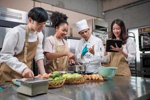 Hobby cuisine course, senior male chef in cook uniform teaches young cooking class students to prepare, mix and stir ingredients for pastry foods, fruit pies in restaurant stainless steel kitchen. photo