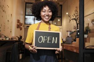 One African American female startup barista stands at casual cafe door, looks at camera, and shows open sign, happy and cheerful smiles with coffee shop service jobs, and new business entrepreneurs. photo