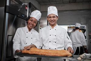 retrato de chefs profesionales con uniforme blanco mirando la cámara con una sonrisa alegre y orgullosa con una bandeja de pan en la cocina. amigo y compañero de la repostería y del oficio diario de panadería fresca. foto