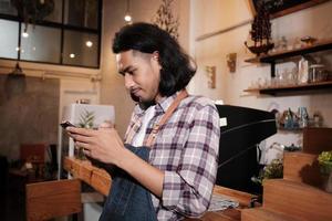 barista asiático de cabello largo y guapo con un delantal se encuentra en un café informal, charla sobre pedidos en línea en la aplicación de teléfono móvil y emprendedor de trabajos de servicio de cafetería. foto