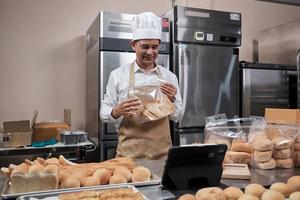 chef asiático senior en delantal, blogger de pequeñas empresas que transmite en vivo a través de tecnología de aplicaciones móviles, promoción de ventas de panadería en línea, pan fresco, pastelería y cocina comercial en la cocina.