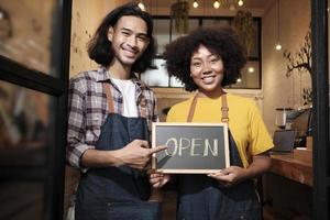 Two young startup barista partners with aprons stand at casual cafe door, letters on board and show open sign, happy and cheerful smiles with coffee shop service jobs, and new business entrepreneurs. photo