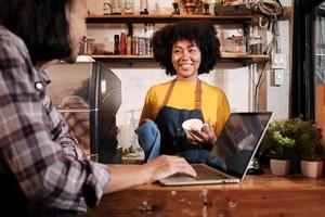 Two cafe business startup partners and friends, African American female, and Thai male baristas talk and cheerful smile together at counter bar of coffee shop, happy service job, and SME entrepreneur. photo