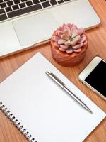 Blank notebook with smartphone, laptop computer and succulent plant in copper pot on wooden table background photo