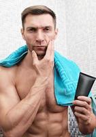 fit toned man holding after shave balm and touching his face. shirtless man in a bathroom with blue towel on shoulders photo