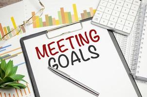 On a chart background magnifier, a pen and a sheet of paper with the text MEETING GOALS photo