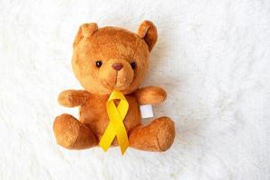 cinta amarilla con muñeca de oso sobre fondo blanco para apoyar la vida y la enfermedad de los niños. mes de concientización sobre el cáncer infantil de septiembre y concepto del día mundial contra el cáncer foto