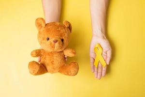 mano sosteniendo una cinta amarilla y una muñeca de oso sobre fondo amarillo para apoyar la vida y la enfermedad de los niños. mes de concientización sobre el cáncer infantil de septiembre y concepto del día mundial contra el cáncer foto