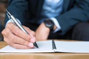 Businessman in suit writing something on notebook in office or cafe, hand of man holding pen with signature on paper report. business concepts photo