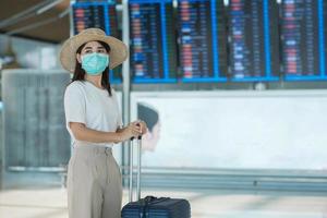 mujer joven con mascarilla con equipaje caminando en el aeropuerto, protección contra la infección por coronavirus, viajera asiática con sombrero. tiempo para viajar, probar e ir y el concepto de dosis de refuerzo de la vacuna foto