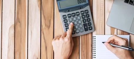 vista superior mujer de negocios escribiendo a mano y calculando con calculadora, bolígrafo, computadora portátil y maceta en el fondo de la mesa de escritorio de oficina de madera. espacio de trabajo u oficina en casa con espacio de copia para el concepto de texto foto