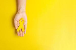 mano sosteniendo una cinta amarilla sobre fondo amarillo para apoyar a las personas que viven y están enfermas. día de la prevención del suicidio, cáncer de sarcoma y concepto del mes de concientización sobre el cáncer infantil foto