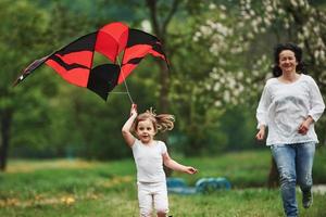 Slower, please. Positive female child and grandmother running with red and black colored kite in hands outdoors photo