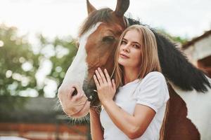 Attractive girl looks straight into the camera. Happy woman with her horse on the ranch at daytime photo