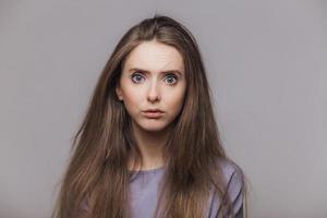 Studio shot of pleasant looking blue eyed female model with dark long hair, has puzzled expression as hears shocking news from interlocutor, isolated over grey background. Facial expressions concept photo