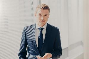 Indoor shot of clever businessman in formal clothes, holds smart phone, prepares for business meeting in office, has work break, stands near window with shutter. Technology and occupation concept photo