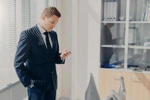 Business, office, people and technology concept. Successful businessman holds smart phone, reads text message from investor, stands in his cabinet, wears formal clothes, connected to wifi at office photo