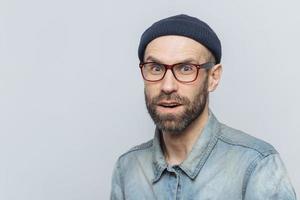 Stylish middle aged male with suprised and curious expression wears spectacles, denim jacket and hat, poses against grey background with copy space for your advertisment or promotional text. photo