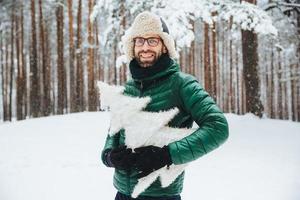 atractivo hombre barbudo con apariencia atractiva, usa anorak verde y sombrero, sostiene un abeto artificial, pasa los fines de semana en un clima helado en el bosque de invierno. concepto de invierno, clima y estilo de vida foto