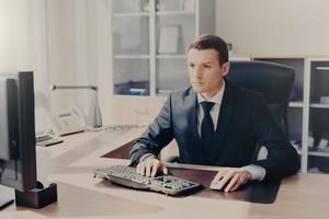 un hombre de negocios serio y concentrado se sienta frente a la computadora en el lugar de trabajo, escribe información, usa traje, prepara un proyecto de negocios para el próximo año, posa en un gabinete espacioso. gente y finanzas foto