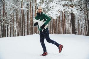 apuesto joven sonriente usa gorro y anorak, pasa tiempo libre al aire libre en el bosque durante el invierno, se prepara para las vacaciones, sostiene un abeto. un tipo barbudo con anteojos admira los paisajes invernales foto