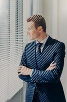 retrato en la cintura de un apuesto financiero masculino mira atentamente en la ventana de la oficina, tiene una expresión pensativa, descansa después de la conferencia, tiene un descanso en el trabajo, mantiene las manos cruzadas, usa ropa elegante foto