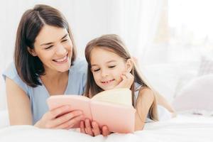 Pretty female mother and her small daughter read interesting book in bedroom, lie on comfortabled bed during morning. Lovely mum reads fairty tale for her little female kid, who is very curious photo