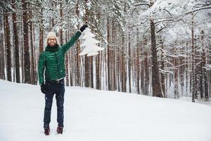 un buen modelo masculino de mediana edad usa una gorra de piel cálida con orejeras y un abrigo acolchado verde, sostiene un abeto artificial blanco, se para en el suelo cubierto de nieve, tiene una expresión feliz foto