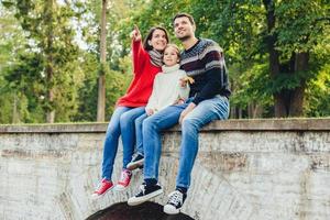 Togetherness and family concept. Friendly father, mother and daughter sit on stone bridge, look from the top, notice beautiful rainbow on sky, indicate on it with fingers, enjoy nature landscapes photo