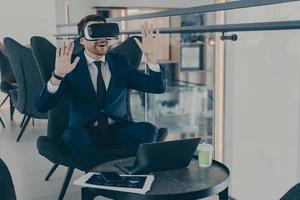 Businessman dressed in formal suit wearing VR goggles while working on laptop in cafe photo