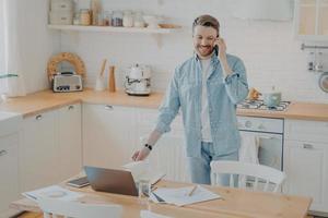 Handsome smiling bearded man speaking on cellphone in kitchen while working remotely photo