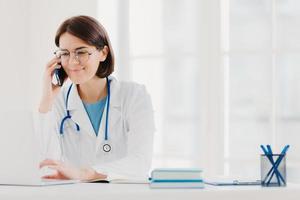 Smiling therapist focused at screen of laptop computer, calls via modern smartphone, wears medical gown, transparent glasses, sits at coworking space, surfes internet. Copy space for your information photo