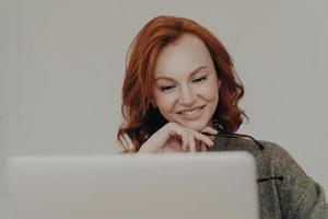 Successful female student with ginger hair and pleasant smile watches interesting tutorial video or webinar, uses laptop computer for e learning, reads information online, keeps hand under chin photo