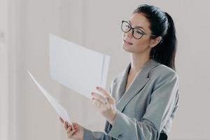 Photo of female financial analyst makes report with recommendations for investments, reads strategy information, poses with paper documents, dressed in formal clothes, wears eyeglasses, stands indoor