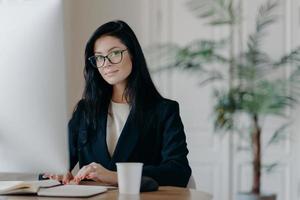 Serious successful female office worker in formal outfit, wears eyeglasses, busy with online work, poses at desktop, works on computer, reads business new or watches tutorial, enjoys working process. photo