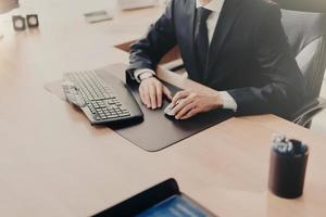 Cropped image of successful male business owner sits at work place in formal suit, uses computer for work, prepares financial report. Male executive works in office alone. People and finances photo