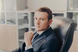 Indoor shot of serious confident businessman poses in chair, poses at cabinet, waits for colleagues to discuss main issues of work. Young male director in formal suit, thinks about new project photo