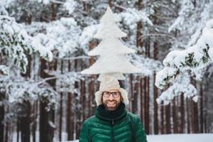 Horizontal shot of smiling male model holds fir tree on head, stands outdor in winter forest, enjoys calmness and tranquil atmosphere. Caucasian unshaven man with cheerful expression photo